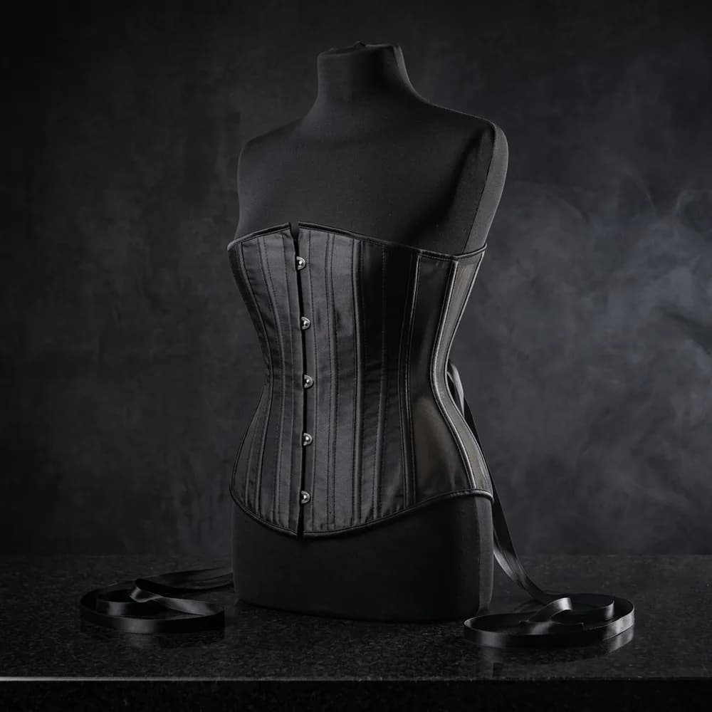 Black underbust corset with sheer mesh side panels and front busk clasps, styled on a dress form